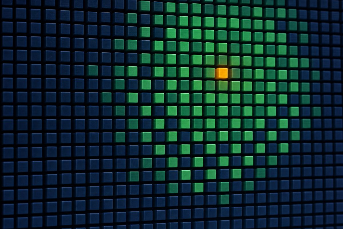 Editorial photograph of a large vertical pegboard grid of matte navy, phosphor-green and amber cubes — a single amber cube sits two-thirds up the grid with a soft circular gradient of phosphor-green around it, as if a search heuristic had radiated outward.